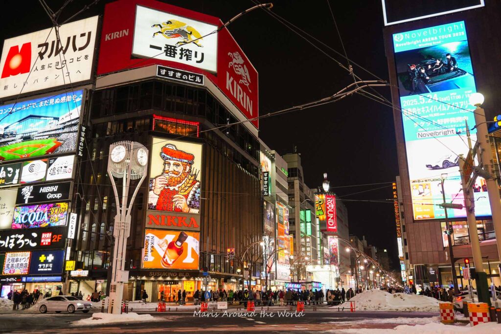 A bustling city street at night, lined with illuminated billboards and snow-covered sidewalks, with a large clock tower in the foreground.